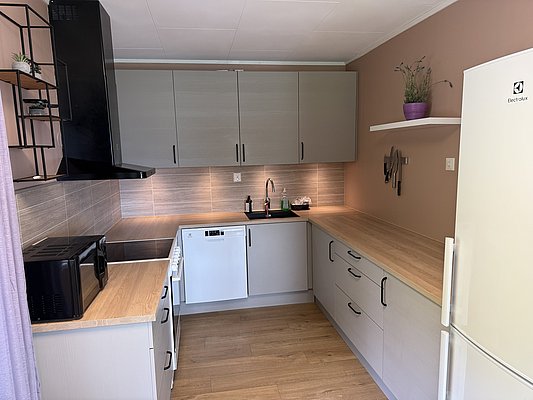 Moderne Küche in Nord-Norwegen mit hellen Holzarbeitsflächen, grauen Schränken und weißem Kühlschrank. Einladendes Ambiente mit Mikrowelle, Spülmaschine und Akzenten in lila. Ideal für Ferienhäuser in idyllischer Umgebung. Perfekt für Angelurlauber.