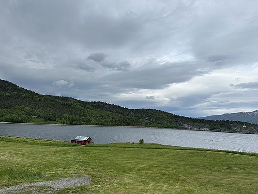 Grüne Wiesen und ruhiges Wasser vor bewaldeten Hügeln und grauem Himmel in Nord-Norwegen. Ein rotes Haus am Seeufer steht im Vordergrund. Ideal für Angelabenteuer und Naturerlebnisse.