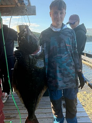 Junger Angler präsentiert riesigen Heilbutt auf Holzsteg in Nord-Norwegen. Im Hintergrund klare Gewässer und bewaldete Hügel. Perfekte Bedingungen für Angelreisen und unvergessliches Meeresangeln.