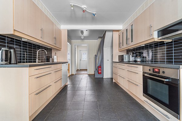 Moderne Küche in Nord-Norwegen: helle Holzschränke, dunkle Bodenfliesen, stilvolle schwarze Fliesenrückwand, integrierter Backofen, Edelstahlspüle, und Wasserkocher. Geräumig und einladend, ideal für Angelurlaube.