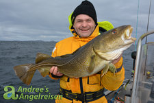 Ein Angler in gelber Jacke und Mütze hält stolz einen großen Kabeljau auf einem Boot vor einer rauen Meereslandschaft. Ein Angler in gelber Jacke und Mütze hält stolz einen großen Kabeljau auf einem Boot vor einer rauen Meereslandschaft.
