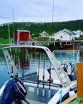 In Nord-Norwegen am Steg liegt ein Motorboot mit Angelruten im Wasser. Im Hintergrund sind bunte Holzhäuser vor grünen Hügeln und schneebedeckten Bergen zu sehen. Perfekt für traumhaften Angelurlaub.