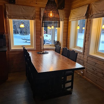 Gemütlicher, holzgetäfelter Innenraum in Nord-Norwegen mit großem Esstisch und stilvoller Lampe, die eine warme Atmosphäre schafft. Die Fenster bieten Ausblick auf verschneite Landschaft, ideal für erholsamen Angelurlaub.
