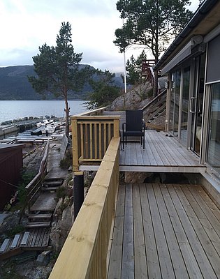 Holzterrasse mit Blick auf norwegischen Fjord, Boote im Wasser, umgeben von Kiefern und Felsen. Ideal für Angelurlaub in Südwest-Norwegen. Entspannung und Abenteuer in wunderschöner Naturkulisse erleben.