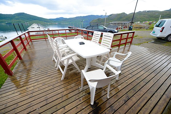 Weitläufige Terrasse mit weißen Gartenmöbeln in Nord-Norwegen. Blick auf Fjord und grüne Berge. Ideal für Angelreisende, die Ruhe und Natur genießen möchten. Perfekte Kulisse für unvergessliche Angelabenteuer.