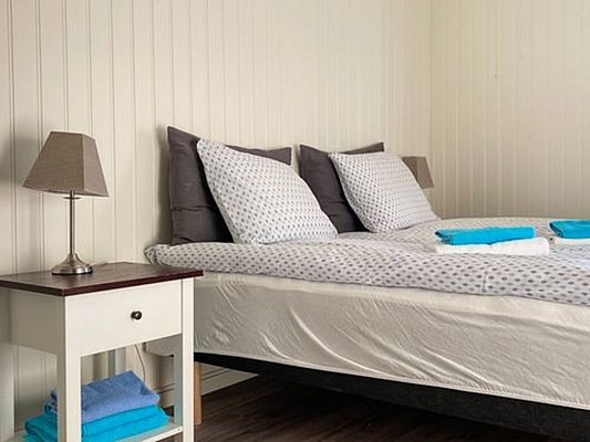 Gemütliches Schlafzimmer in Nord-Norwegen mit Doppelbett, weißer Holzverkleidung und Nachttischlampe. Ideal für erholsame Angelreisen und entspannende Nächte. Frische Handtücher in Blau und Weiß ergänzen die ruhige Atmosphäre.