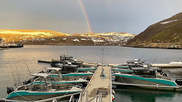Ein malerischer Hafen in Nord-Norwegen bei Abendlicht. Mehrere Anglerboote liegen vertäut, während ein Regenbogen über schneebedeckten Hügeln erscheint. Perfekter Ausgangspunkt für Angelreisen im klaren, ruhigen Wasser.