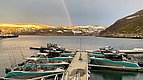 Ein malerischer Hafen in Nord-Norwegen bei Abendlicht. Mehrere Anglerboote liegen vertäut, während ein Regenbogen über schneebedeckten Hügeln erscheint. Perfekter Ausgangspunkt für Angelreisen im klaren, ruhigen Wasser.
