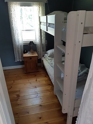 Das Bild zeigt ein kleines Schlafzimmer mit Etagenbett, Holzfußboden, einem Fenster mit weißen Gardinen und einem Nachttisch.