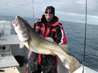 Eine Person in dicker Outdoor-Kleidung hält einen großen Kabeljau auf einem Boot mit dem Meer im Hintergrund.
