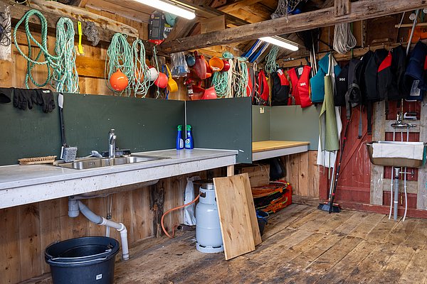 Rustikale Werkstatt in Hitra, Norwegen: Angelzubehör, bunte Rettungswesten und Seile an Holzwänden. Edelstahlspüle, Arbeitsflächen und robuste Ausstattung schaffen eine praktische Umgebung für Angler. Perfekt für Vorbereitungen und Ausrüstungspflege vor dem Meeresangeln.
