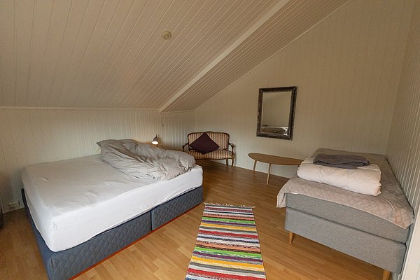 Gemütliches Schlafzimmer in Nord-Norwegen mit schräger Holzdecke, Doppelbett, Sofa, modernem Spiegel und buntem Teppich. Ideal für entspannten Angelurlaub. Helle, einladende Atmosphäre bietet Komfort nach erlebnisreichen Angeltouren. Perfekt für Erholungssuchende.