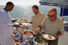 Drei Männer bedienen sich an einem Buffet auf einem Boot mit Blick auf einen See unter blauem Himmel.