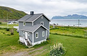 Graues Holzhaus in Nord-Norwegen vor beeindruckender Küstenlandschaft. Im Hintergrund Meer und Berge, umgeben von grüner Wiese. Perfekt für einen idyllischen Angelurlaub in unberührter Natur.
