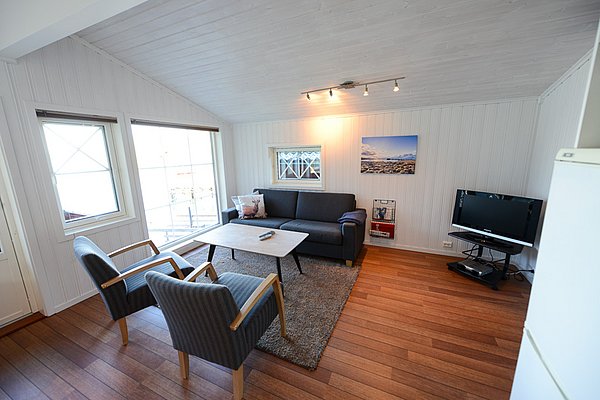 Gemütliches Wohnzimmer in Nord-Norwegen, schlicht skandinavisch eingerichtet. Helle Wände, zwei Fenster mit Tageslicht, Sofa, zwei Sessel, Tisch, Fernseher. Ideal für Entspannung nach einem erfolgreichen Angelurlaub.