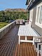 Große Holzterrasse mit weißem Tisch und Sitzmöbeln, umgeben von üppigem Grün und Bergen in Nordland, Norwegen. Perfekte Kulisse für Angelurlaub und naturnahe Erholung.