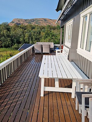 Große Holzterrasse mit weißem Tisch und Sitzmöbeln, umgeben von üppigem Grün und Bergen in Nordland, Norwegen. Perfekte Kulisse für Angelurlaub und naturnahe Erholung.