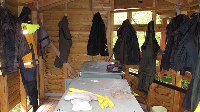 Holzhütte in Hitra, Norwegen, mit hängenden Regenjacken und Arbeitskleidung. Auf dem Tisch liegen Handschuhe und Ausrüstung. Perfekt für Angler, die Schutz und Komfort suchen. Abenteuerliches Angelreiseziel in natürlicher Umgebung.
