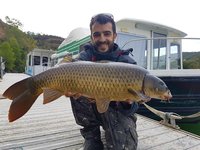 Ein Angler posiert auf einem Steg vor einem Boot und hält einen großen Karpfen in den Händen.