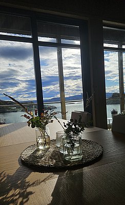 Blick aus einem norwegischen Ferienhaus auf die idyllische Küstenlandschaft von Hitra. Durch große Fenster strömt Tageslicht, das den traumhaften Meerblick und die stilvollen Glaselemente auf einem Holztisch betont. Perfekte Kulisse für einen Angelurlaub.