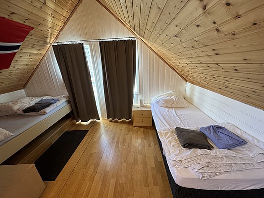 Gemütliches Schlafzimmer in Hitra, Norwegen, mit Holzwänden und zwei Einzelbetten. Fenster mit Vorhängen, Holzboden und norwegischer Flagge. Perfekt für erholsame Nächte nach erfolgreichen Angelabenteuern.