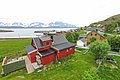 Ein charmantes rotes Haus steht vor der Kulisse verschneiter Berge und dem Meer in Nord-Norwegen. Umgeben von grünen Wiesen bietet es eine idyllische Atmosphäre für Angelurlaub und Naturerlebnisse.