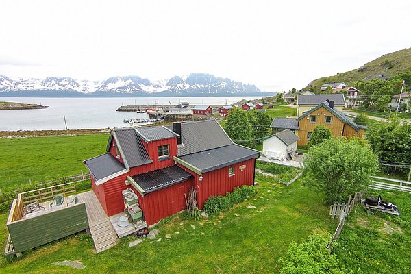 Ein charmantes rotes Haus steht vor der Kulisse verschneiter Berge und dem Meer in Nord-Norwegen. Umgeben von grünen Wiesen bietet es eine idyllische Atmosphäre für Angelurlaub und Naturerlebnisse.