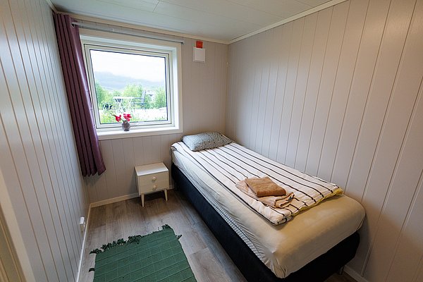 Kompaktes Schlafzimmer in Nord-Norwegen: Einzelbett, gestreifte Bettwäsche, Fensterblick auf grünen Garten, Bergkulisse. Ideal für Anglerurlaub. Helle Holzwände, ländlich-modern eingerichtet, komfortable Erholung nach erfolgreichem Angelabenteuer.