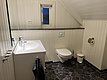 Ein modernes Badezimmer in Nord-Norwegen mit weißer Holzverkleidung, Dachschräge und kleinem Fenster. Elegante Armaturen am Waschbecken, grauer Handtuchhalter, WC und schwarzer Fliesenboden. Ideal für komfortable Angelunterkünfte.