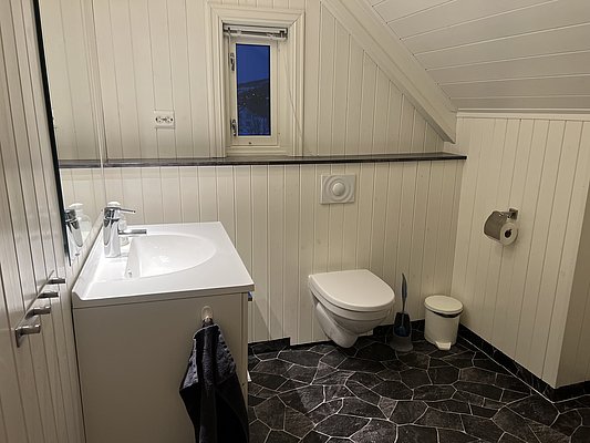 Ein modernes Badezimmer in Nord-Norwegen mit weißer Holzverkleidung, Dachschräge und kleinem Fenster. Elegante Armaturen am Waschbecken, grauer Handtuchhalter, WC und schwarzer Fliesenboden. Ideal für komfortable Angelunterkünfte.