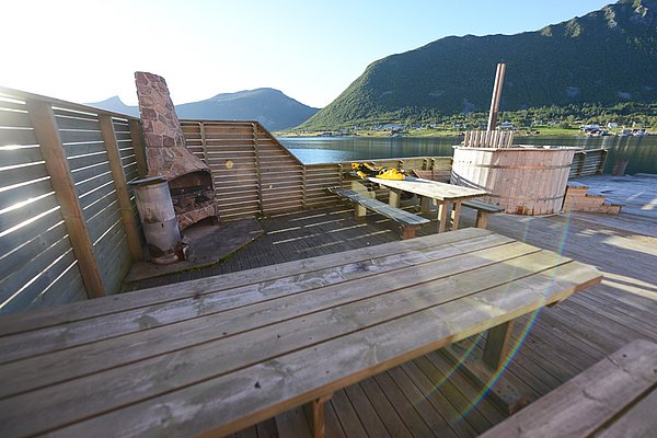 Holzterrasse mit Grill und Hot Tub in Nord-Norwegen, Blick auf malerischen Fjord und Berge, ideal für entspannte Angelurlaub-Momente. Perfekte Kulisse für Outdoor-Abenteuer und Erholung inmitten atemberaubender Natur.