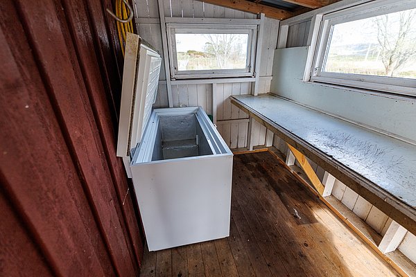 Kleiner, rustikaler Raum mit weißer Gefriertruhe und Arbeitstisch, ideal zur Fischverarbeitung nach erfolgreichen Angelreisen in der Region Hitra, Norwegen. Perfekt für Angler auf der Suche nach Abenteuer und Komfort.