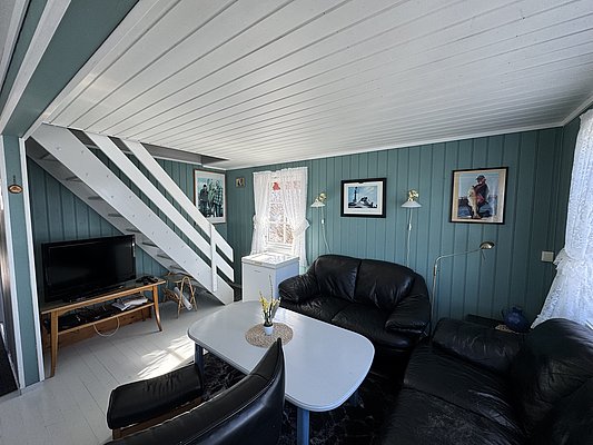 Gemütliches Wohnzimmer in Hitra, Norwegen: Moderne Einrichtung mit schwarzen Ledersofas, Holztisch und Treppe. Helle Wände mit Fischbildern schaffen maritimes Flair. Ideal für entspannenden Angelurlaub.