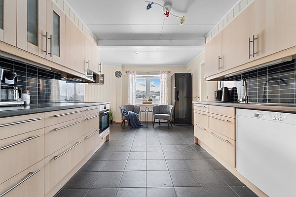 Moderne, helle Küche in Nord-Norwegen mit Holzschränken und schwarzen Fliesen. Am Fenster zwei Sessel mit Tisch, bieten Aussicht auf verschneite Landschaft. Ideal für Angelreisende als gemütlicher Rückzugsort nach einem erlebnisreichen Tag.