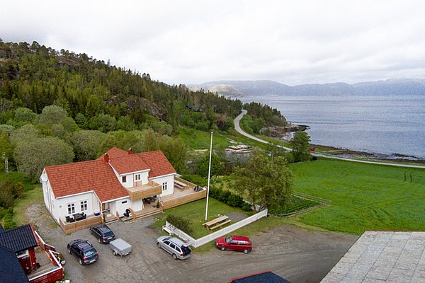 Die Aufnahme zeigt ein gemütliches rotes Haus in Hitra, Norwegen, umgeben von üppigem Grün, mit Blick aufs Meer. Perfekt für Angelreisen, bietet die Umgebung Ruhe und Naturerlebnis pur.
