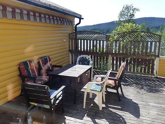 Eine sonnige Terrasse vor einem gelben Haus mit mehreren bunten Stühlen und einem Tisch, umgeben von grünem Wald und mit Blick auf einen See.