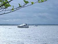 Ein Boot fährt auf einem ruhigen See unter einem bewölkten Himmel, während ein Ast mit frischen grünen Blättern das Bild im Vordergrund ergänzt.
