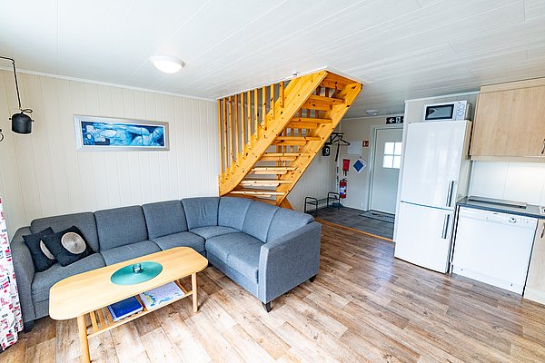 Gemütliches Wohnzimmer in Nord-Norwegen mit grauem Ecksofa, Holztisch und moderner Küche. Perfekte Unterkunft für Angelreisen im skandinavischen Stil. Helle, einladende Atmosphäre mit Holztreppe und mariner Wanddekoration.