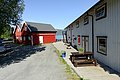 Rotes und weißes Gebäude in Nordland, Norwegen, mit Blick auf einen klaren Fjord. Holzveranda mit Sitzbereich, umgeben von Bäumen. Perfekte Kulisse für einen entspannten Angelurlaub im skandinavischen Stil.
