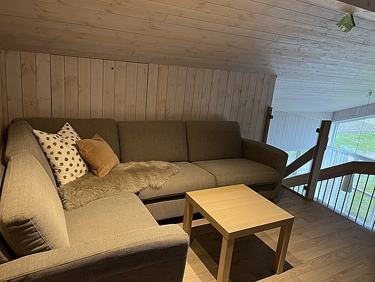 Gemütliches Wohnzimmer in einer Hütte in Hitra, Norwegen. Holzwände, bequeme Eckcouch mit Kissen und Fell, kleiner Holztisch. Ideal für entspannten Angelurlaub und erholsame Abende nach einem erfolgreichen Tag am Meer.