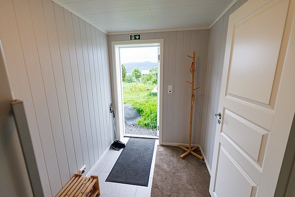 Hausflur in Nord-Norwegen, Blick ins Grüne durch offene Tür, mit Holzgarderobe und Fußmatte. Ideal für Angelurlauber, bietet schnellen Zugang zur Natur. Entspannte, lichtdurchflutete Atmosphäre in skandinavischem Design.