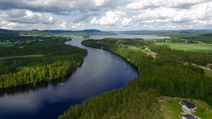 Luftaufnahme von Hälsingland, Schweden: Fluss schlängelt sich durch grüne Wälder und Felder unter dramatischem Himmel. Ideal für Angelurlaub und Naturerlebnisse. Herrliche Landschaft und Ruhe für Outdoor-Fans und Abenteurer.