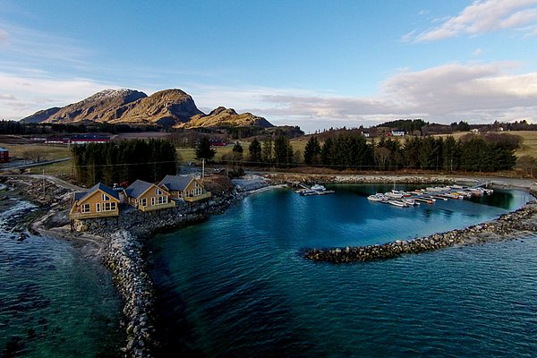 Norwegisches Nordland: Malerische Küstenlandschaft mit gelben Holzhäusern, Booten im klaren Wasser und majestätischen Bergen im Hintergrund unter blauem Himmel. Perfekt für Angelreisen und Naturerlebnisse.