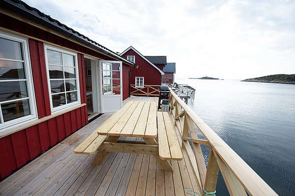 Ein rotes, am Wasser gelegenes Ferienhaus mit Terrasse, Holztisch und weiter Aussicht auf das Meer.