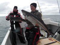 Zwei Angler in warmen Jacken stehen auf einem Boot und halten stolz zwei große Fische, während sie auf dem offenen Meer sind.