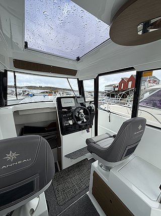 Modernes Innenraumfoto eines Angelbootes in Süd-Norwegen. Helle Kabine mit Steuerkonsole und komfortablen Sitzen. Durch das Dachfenster fällt Regen, Hafenkulisse im Hintergrund. Ideal für Angelabenteuer in Norwegen.