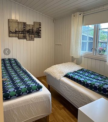 Gemütliches Schlafzimmer auf Hitra, Norwegen: Zwei Einzelbetten mit geometrischen Decken, Holzboden, weiß getäfelte Wände. Großes Fenster mit Blick auf die Umgebung. Perfekte Unterkunft für Angelreisen und Naturliebhaber.