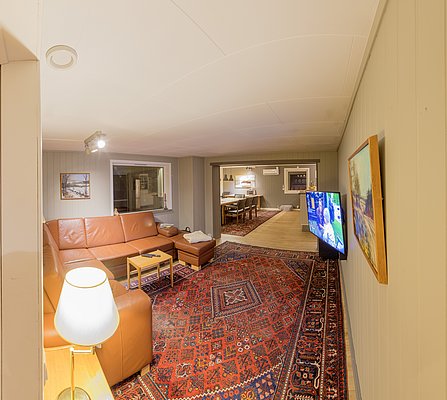 Gemütliches Wohnzimmer in Nordland, Norwegen mit rotem Teppich, beiger Ledercouch und Flachbild-TV. Blick in den Essbereich, helle Wände, Lampenlicht. Perfekt für Erholung nach einem spannenden Angeltag. Ideal für Angelurlaube.