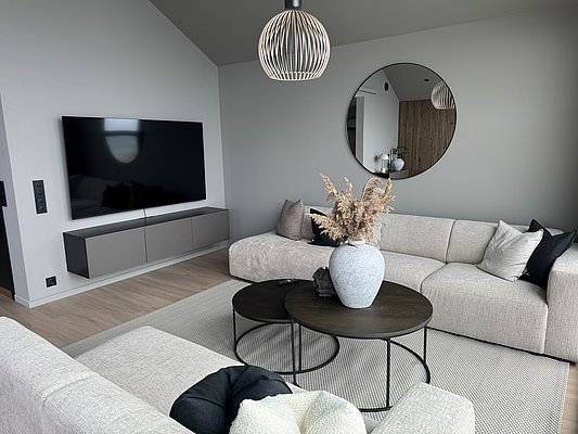 Modernes, stilvolles Wohnzimmer in Südwest-Norwegen: Helle, beige Sofas, runder Couchtisch mit Dekoration, großer Fernseher und runder Spiegel an grauen Wänden. Perfekt für gemütliche Entspannung im skandinavischen Design. Keywords: Skandinavisches Wohnzimmer, modernes Interieur.