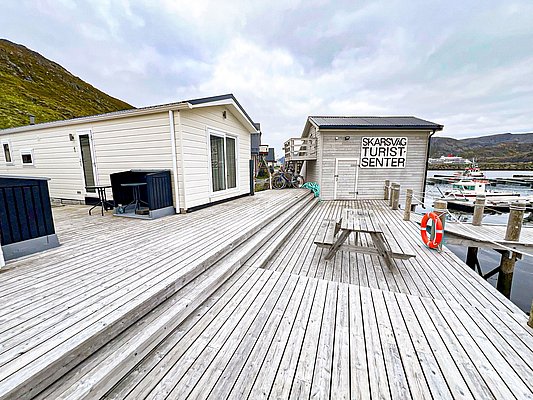 Holzterrasse am Skarsvåg Touristensenter in Nordnorwegen mit Blick auf den Hafen. Traditionelle Holzhäuser bieten gemütliche Unterkünfte, ideal für Angelurlaub. Fjordlandschaft im Hintergrund verstärkt das Naturerlebnis. Perfekte Kombination aus Ruhe und Abenteuer.